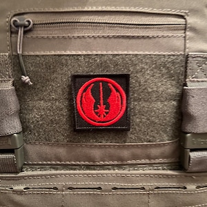 Op de afbeelding: Een grijze tactische vest met een rood en zwart patch met een gestileerd symbool. De patch is bevestigd aan de voorkant van de vest.
