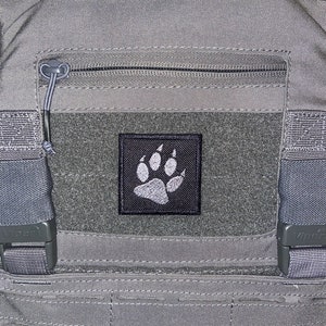 May include: A grey tactical vest with a black patch featuring a white paw print. The vest has a zipped pocket and adjustable straps. The vest also has a small Union Jack patch and the number "387" printed on the top right corner.