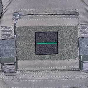 May include: A grey tactical vest with a black patch featuring a thin green line. The vest has two adjustable straps with black buckles.