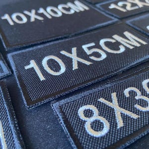 Może przedstawiać: Zbliżenie na czarne łatki z tkaniny z białym haftowanym tekstem. Łatki mają różne rozmiary, w tym 10x10cm, 10x5cm i 8x3cm.