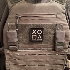 May include: Grey tactical vest with a front zip, Velcro patches, and a grid of horizontal straps. A black square patch displays the letters XO and geometric shapes. The vest has adjustable straps and the word "Viper" is visible.