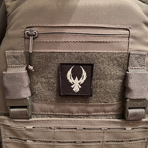 Op de afbeelding: Een grijze tactische vest met een centraal embleem met een wit embleem. De vest heeft riemen, gespen en een zak met ritssluiting. Het ontwerp benadrukt functionaliteit en duurzaamheid.