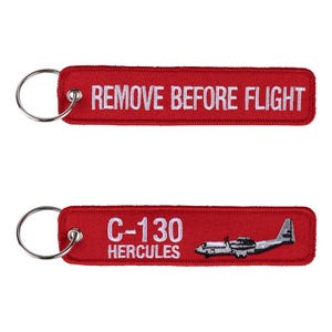 Pode incluir: Dois porta-chaves retangulares de tecido bordado vermelho com argolas de metal prateado. O porta-chaves superior apresenta o texto branco REMOVE BEFORE FLIGHT. O inferior exibe o texto branco C-130 HERCULES e um gráfico de um avião.