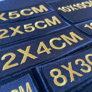 May include: Close-up of several black fabric patches with gold embroidered text indicating different sizes in centimetres. The patches include 5x5cm, 12x5cm, 12x4cm, 8x3cm, 10x10cm, and 10x5cm.