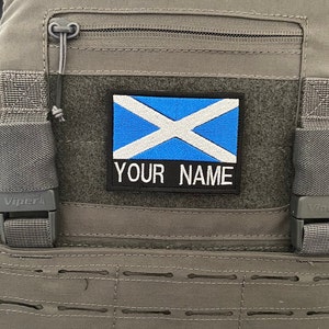 Op de afbeelding: Een grijze tactische vest met een blauw-witte Schotse vlag patch met de tekst "YOUR NAME" erop.