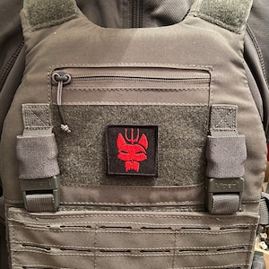 May include: A grey tactical vest with a black patch featuring a red stylised wolf with a trident on its head. The vest has a zipped pocket and adjustable straps.