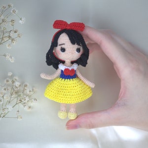 May include: A handmade crochet doll of a character with black hair, a red bow, and a yellow dress. The doll has a white collar with a red heart and blue accents. The doll is approximately 4 inches tall.