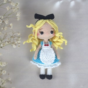 May include: A crocheted doll with blonde hair, a blue and white dress, and a black bow. The doll is wearing a white apron with a red bow.