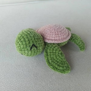May include: A green and pink crocheted turtle toy with a closed eye expression. The turtle is lying on its side with its head resting on the surface.