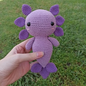 May include: A purple crocheted axolotl plush toy with black eyes and a pink blush. The toy is being held by a hand.