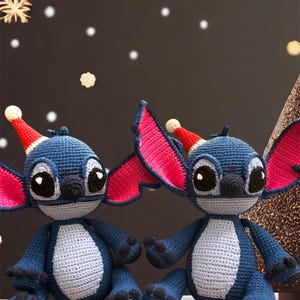 May include: Two blue and gray crocheted Stitch characters wearing red and white Santa hats. The characters have large black eyes and are sitting on a white surface.