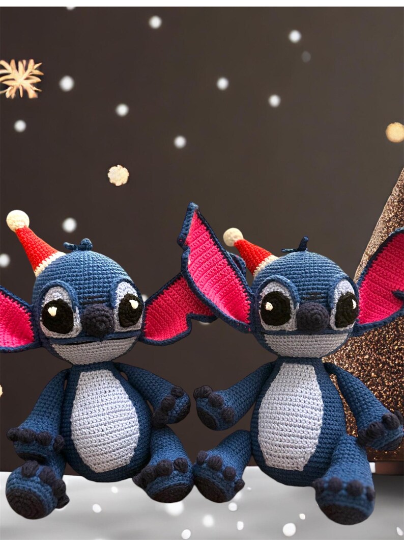 May include: Two blue and gray crocheted Stitch characters wearing red and white Santa hats. The characters have large black eyes and are sitting on a white surface.