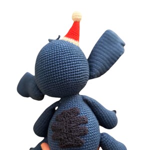 May include: Blue crocheted Stitch plush toy wearing a red and white Santa hat. The toy has a black four-leaf clover patch on its back.