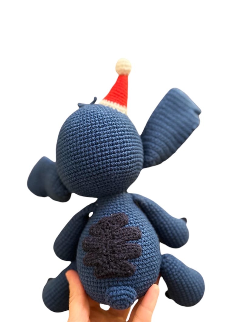 May include: Blue crocheted Stitch plush toy wearing a red and white Santa hat. The toy has a black four-leaf clover patch on its back.