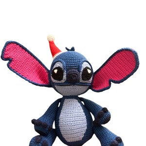 May include: A blue and pink crocheted Stitch toy wearing a red Santa hat. The toy is sitting with its arms crossed and has a friendly expression.