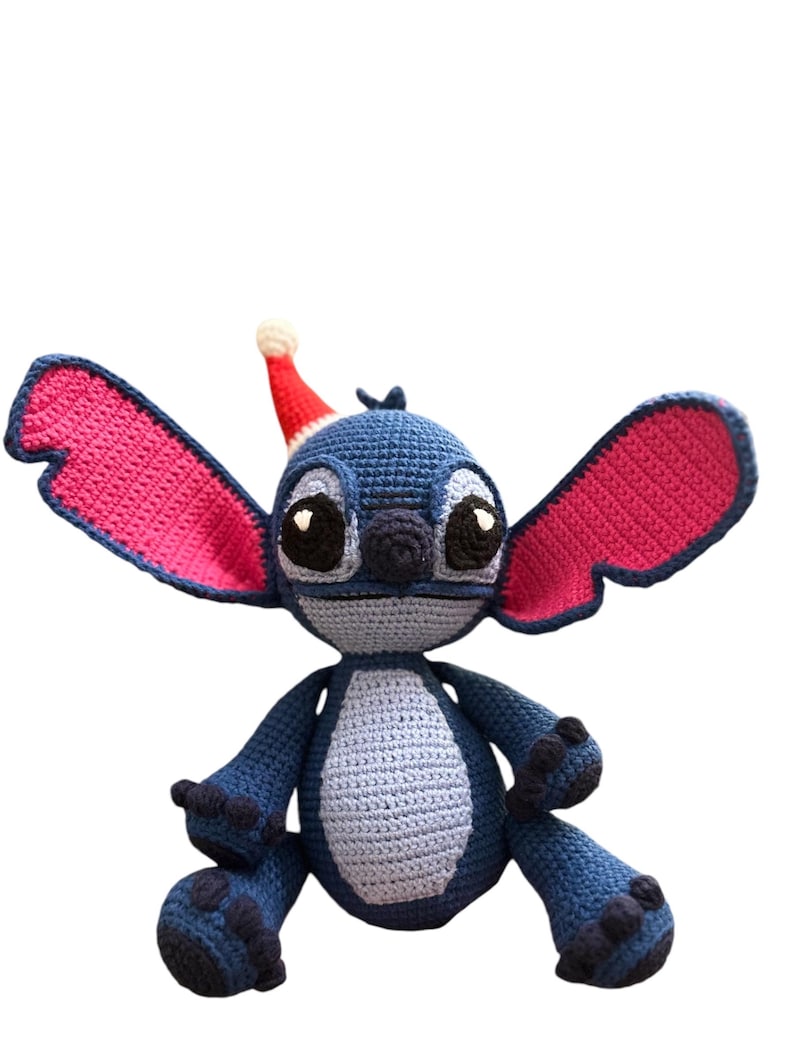 May include: A blue and pink crocheted Stitch toy wearing a red Santa hat. The toy is sitting with its arms crossed and has a friendly expression.