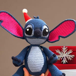 May include: Blue crocheted Stitch character wearing a red Santa hat. The character has large pink ears and is sitting on a red box with a snowflake design.