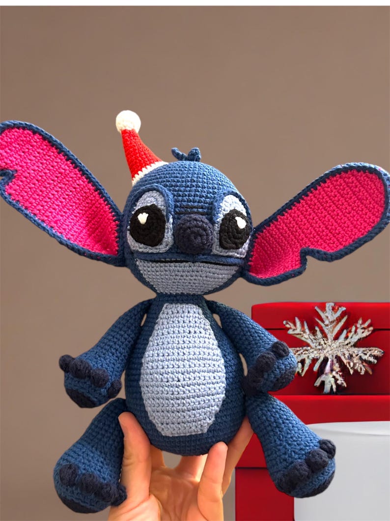 May include: Blue crocheted Stitch character wearing a red Santa hat. The character has large pink ears and is sitting on a red box with a snowflake design.