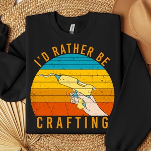 Peut inclure: Sweat-shirt noir avec un graphique de pistolet à colle de style vintage et le texte "I'd rather be crafting".