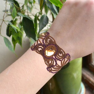 以下が含まれることがあります： 中央に金色の丸い宝石が付いた、茶色と紫のマクラメブレスレットです。