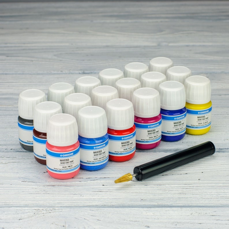 Puede incluir: Un conjunto de 18 peque&ntilde;os frascos de pintura, cada uno con una tapa blanca y un color diferente. Los frascos est&aacute;n dispuestos en semic&iacute;rculo sobre una superficie de madera gris claro. Un aplicador de pintura negro y plateado se encuentra en primer plano.