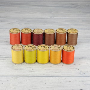 May include: Twelve spools of thread in various shades of orange, yellow, and brown. The spools are labeled with the number "632" and the name of the color in French.