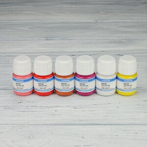 Puede incluir: Seis peque&ntilde;os frascos de pintura MaxEdge Basic Edge Paint en seis colores diferentes: rosa, rojo, marr&oacute;n, magenta, blanco y amarillo. Los frascos est&aacute;n alineados en una superficie de madera clara.