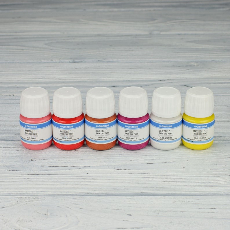 Puede incluir: Seis peque&ntilde;os frascos de pintura MaxEdge Basic Edge Paint en seis colores diferentes: rosa, rojo, marr&oacute;n, magenta, blanco y amarillo. Los frascos est&aacute;n alineados en una superficie de madera clara.