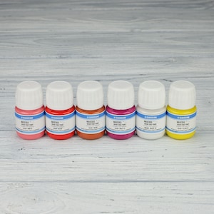 Puede incluir: Seis peque&ntilde;os frascos de pintura MaxEdge Basic Edge Paint en seis colores diferentes: rosa, rojo, naranja, morado, blanco y amarillo. Los frascos est&aacute;n alineados en una fila sobre una superficie de madera clara.