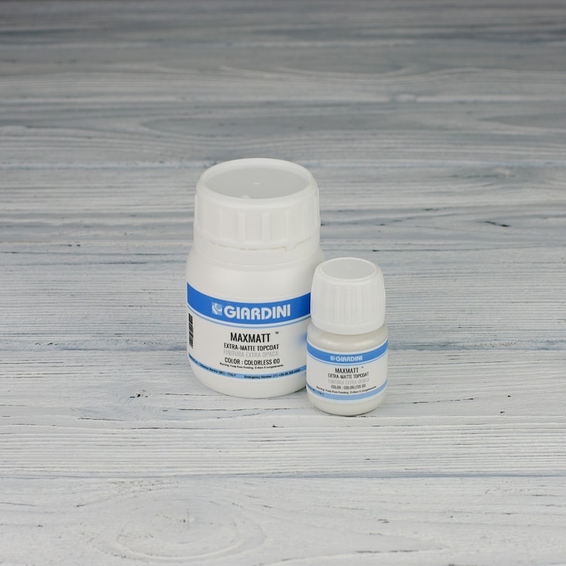 May include: Two bottles of Giardini Maxmatt Extra Matte Topcoat, one large and one small, on a white wood surface. The larger bottle is labelled "Maxmatt Extra Matte Topcoat" and "Color: Colorless 00". The smaller bottle is labelled "Maxmatt Extra Matte Topcoat".