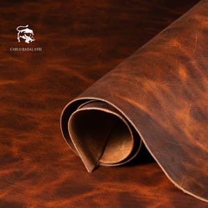 May include: A rolled sheet of distressed brown leather. The leather has a rich, textured appearance with variations in color, ranging from dark brown to lighter tan hues. The Carlo Badalassi logo is visible in the upper left corner.