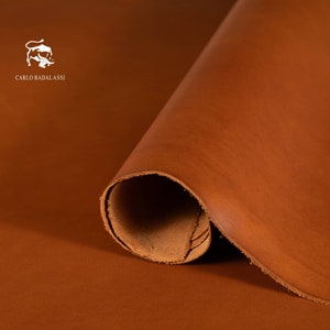 May include: A rolled sheet of brown leather with a smooth surface. The leather is a warm, earthy tone, with a slight sheen. The brand name "CARLO BADALASSI" is printed in white on the upper left corner, with a bull logo.