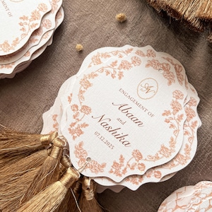 May include: A stack of off-white, scalloped-edge tags with a floral border and the words "Engagement of Abaan and Nashika 07.12.2025" in a brown script. Each tag is adorned with a gold tassel.