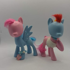 Puede incluir: Dos figuras de ponis de juguete. Uno es azul claro con pelo y alas rosas, y una pulsera con un dije de flor. El otro es azul claro con una cresta roja y cuerpo rosa. Ambos tienen collares de cuentas y colas rosas.