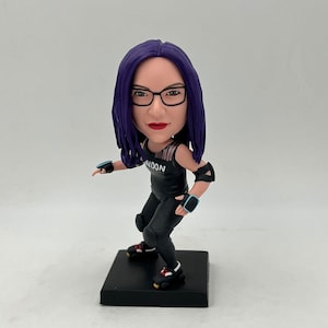 Può includere: Una statuetta con i capelli viola che indossa pattini a rotelle neri, pantaloni neri e una canotta nera con la scritta "NOON". La statuetta indossa ginocchiere e gomitiere. La statuetta è in posizione di pattinaggio con una gamba tesa in avanti e l'altra piegata.