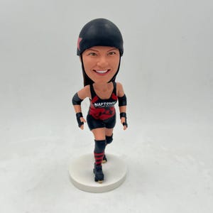 Può includere: Una statuetta bobblehead personalizzata di una giocatrice di roller derby che indossa un casco nero, una maglia rosso e nera con il testo "NAPTOWN ROLLER GIRLS" e ginocchiere nere e rosse. La statuetta è in piedi su una base bianca.