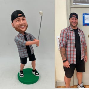 Könnte beinhalten: Eine individuelle Wackelkopf-Figur, die eine Person mit schwarzer Baseballkappe, einem grau-roten karierten Hemd über einem schwarzen T-Shirt und schwarzen Shorts zeigt. Die Figur hält einen Golfschläger und steht auf einem grünen Sockel.
