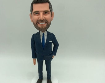 May include: A custom bobblehead figurine featuring a person with short dark hair and a beard, wearing a navy blue suit, a white collared shirt, and a solid blue necktie. The figure stands on a circular white base with a hand in a pocket.