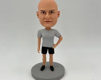 May include: A bald bobblehead figurine featuring a person with a light skin tone, wearing a plain light grey short-sleeved t-shirt, black shorts, and black shoes. The figure stands on a circular grey base, with one hand resting on the hip.