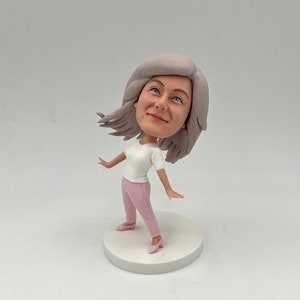 May include: A hand-painted figurine of a woman with gray hair, wearing a white shirt and pink pants. She is standing on a white base.