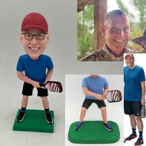 May include: A custom bobblehead figurine depicting a person wearing a red cap, black-rimmed glasses, a blue t-shirt, and black shorts, holding a white and red pickleball paddle while standing on a rectangular green base, modelled after a photograph.