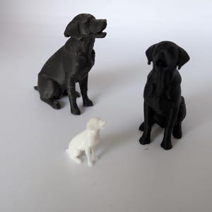 May include: Two black dog figurines and one smaller white dog figurine are positioned in a group. Each dog figurine is depicted in a seated position with ears folded down and heads tilted slightly upward, showing a textured surface finish.