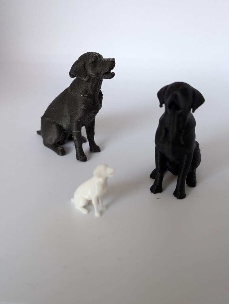 May include: Two black dog figurines and one smaller white dog figurine are positioned in a group. Each dog figurine is depicted in a seated position with ears folded down and heads tilted slightly upward, showing a textured surface finish.