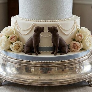 Two black 3D printed labrador retriever cake topper facing each other on a beautiful cake, that is placed on a silver base. Also two glasses are visible on the table.