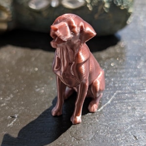 May include: A small, metallic copper-colored figurine of a sitting dog with its mouth slightly open. The sculpture features visible 3D-printed layer lines across its surface, capturing the canine in a seated position with ears folded downward.