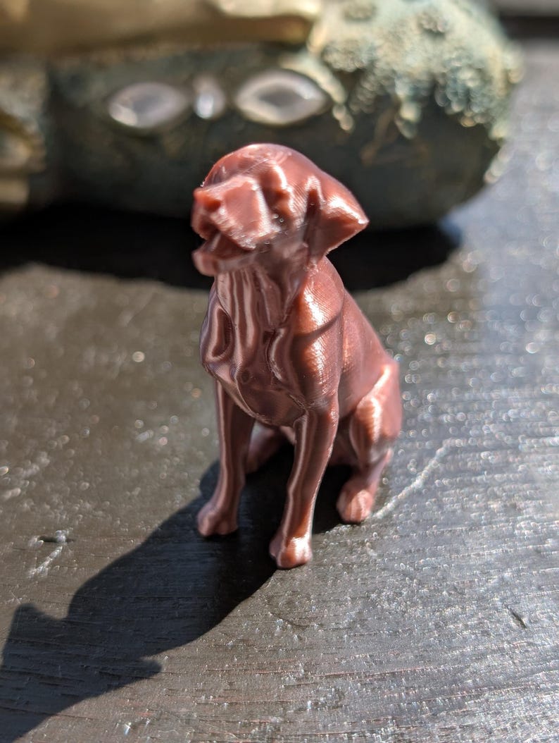 May include: A small, metallic copper-colored figurine of a sitting dog with its mouth slightly open. The sculpture features visible 3D-printed layer lines across its surface, capturing the canine in a seated position with ears folded downward.