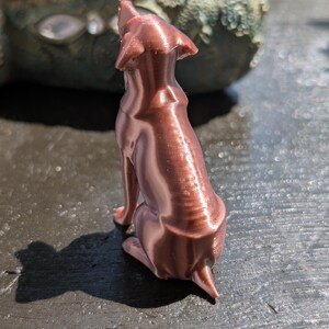May include: A small, metallic copper-colored figurine of a dog in a seated position, viewed from behind. The surface of the dog sculpture displays visible horizontal 3D printing layers, with pointed ears and a short tail clearly defined in the design.