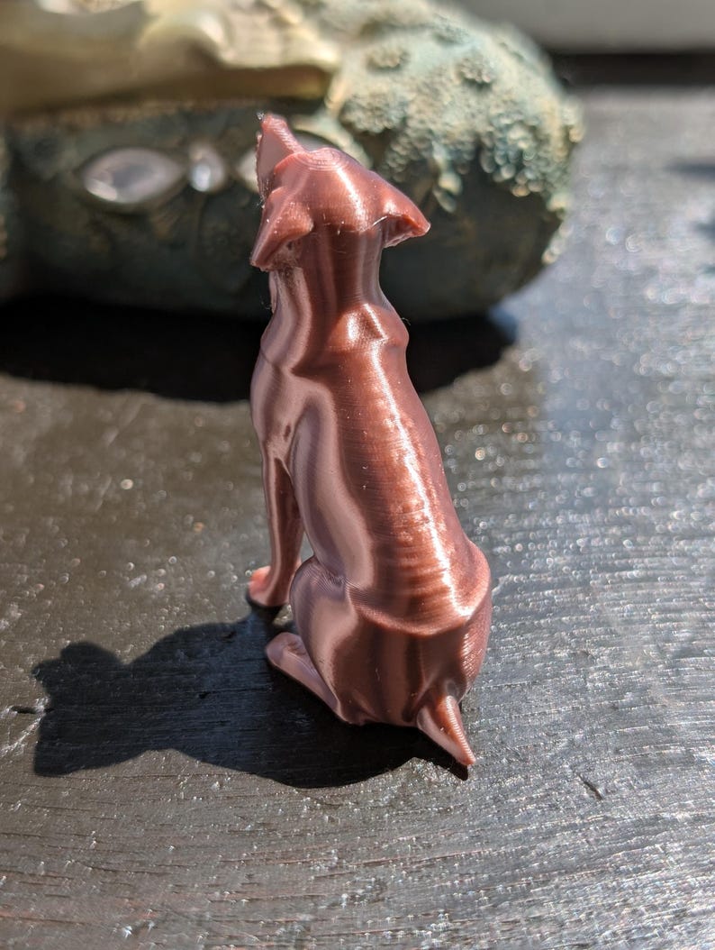 May include: A small, metallic copper-colored figurine of a dog in a seated position, viewed from behind. The surface of the dog sculpture displays visible horizontal 3D printing layers, with pointed ears and a short tail clearly defined in the design.