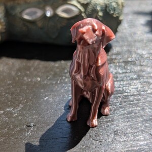 May include: A small, metallic copper-colored figurine of a dog in a sitting position. The figure displays visible 3D-printed layer lines across its surface, with a smooth, reflective finish that catches the light, showing the dog's ears, snout, and body.