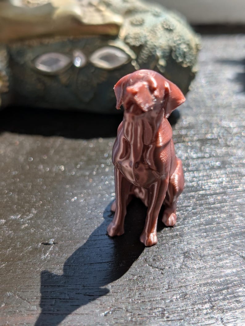 May include: A small, metallic copper-colored figurine of a dog in a sitting position. The figure displays visible 3D-printed layer lines across its surface, with a smooth, reflective finish that catches the light, showing the dog's ears, snout, and body.